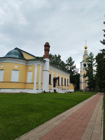 In the city of Vasil'ki, Russia, a low-angle shot captures an outdoor scene featuring a yellow building with a green domed roof on the left and a tall, white and yellow bell tower on the right. The yellow building has white decorative trim and boarded-up windows. A brick chimney rises from the roof of the yellow building. The bell tower features clocks and arches. Both structures are surrounded by a lush green lawn. A paved walkway, composed of red and tan bricks, extends from the foreground into the distance, angling towards the right side of the frame. Tall trees are visible behind the bell tower and to the right of the yellow building. The sky is overcast and grey.