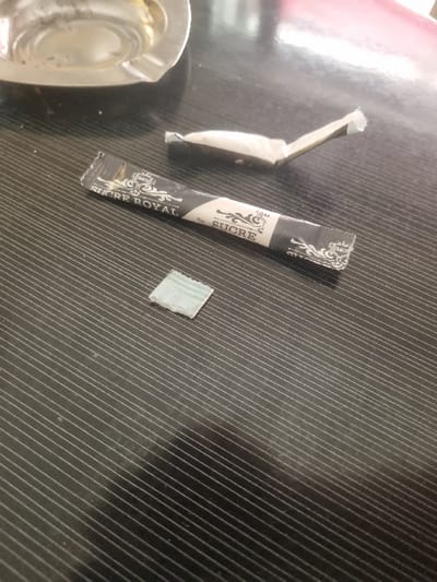 The image shows a black and white sugar packet with the words "Sucre Royal" printed on it, lying open on a dark, lined surface. There is a small, white square object lying beside the packet. In the top left corner of the image, there is a silver ashtray. This scene is located at 36.838 latitude and 10.128 longitude, which is in the city of Tunis, Tunisia. There is no indication of any people or actions taking place in the image.