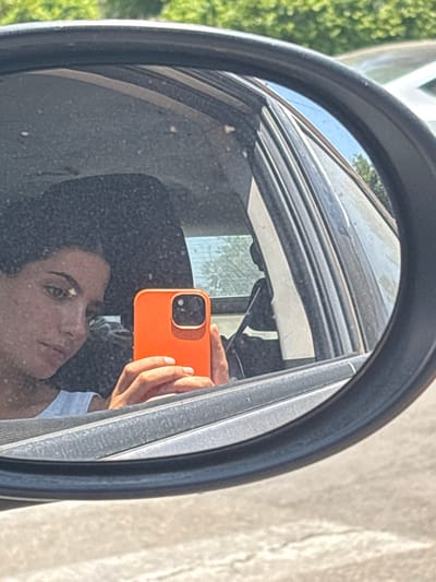 A person is reflected in a car's side mirror, holding an orange smartphone up to take a selfie. The mirror is dirty, with visible smudges and spots. The car's interior is dark, and the window frame is visible above the person's head. Outside the car, there are trees and a building, both slightly blurred in the background. The mirror's edge frames the scene in black.