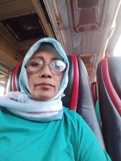 The image is a selfie taken from a bus, with the passenger seated in a black and red seat. The passenger is wearing a teal shirt and a light blue hijab with a patterned scarf. The passenger is also wearing glasses. The bus interior is visible, including the ceiling with ventilation and overhead compartments, and rows of seats. The selfie captures the passenger's face from the chest up. The image appears to be taken during daylight. The location is identified as Jekulo, Indonesia.
