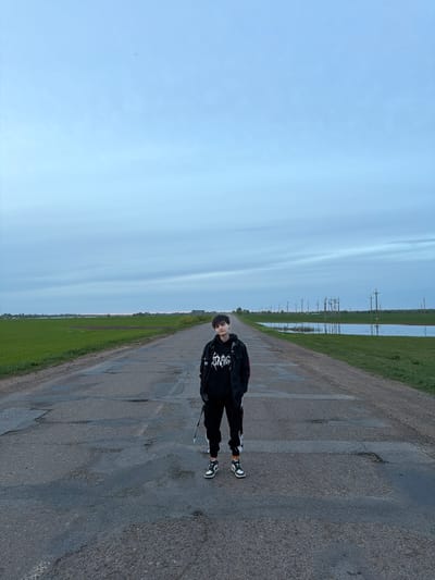 Azyakul', Russia: A person stands on an asphalt road. To the right of the road is green grass. To the left of the road is a field and a blue sky. Power lines can be seen in the distance.