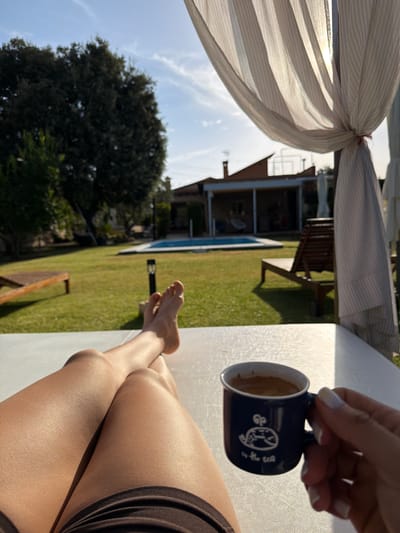A person's legs and feet are visible in the foreground, suggesting they are lounging outdoors. They are holding a dark blue mug with white text and a drawing, possibly enjoying a beverage. The setting appears to be a garden with a swimming pool and a house in the background. Sunlight indicates it is daytime, likely morning or afternoon. The overall mood seems relaxed and peaceful.