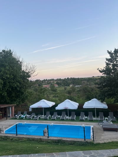 The image depicts an outdoor swimming pool area at dusk, situated in the village of General Kiselovo, Bulgaria. The pool is rectangular and filled with clear blue water. Around the pool, there are several lounge chairs, some with white cushions and others with grey. Three white umbrellas stand near the loungers; one displays the Coca-Cola logo, and another has Cyrillic text, likely a brand name. To the left of the pool area, a small structure with a thatched roof appears to be a bar or changing room, with a Coca-Cola branded refrigerator visible inside. A black metal fence encloses the pool area. Beyond the pool, a gently sloping landscape is visible, dotted with houses and trees, characteristic of a rural Bulgarian setting. The sky above displays a soft gradient of blue and pink, suggesting the end of daylight. Green grass covers the foreground and areas around the pool.