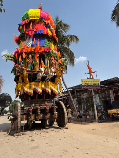 A vertical shot shows a decorated temple chariot in Anegundi, India. The chariot is adorned with colorful fabrics, flags, and garlands. It is elaborately decorated with carvings and statues.

The chariot is positioned on a concrete surface. The backdrop features a building with a sign reading "Sri Hara-Hara Mahadeva Decorator," suggesting a decorator or artisan related to religious events. The sign includes contact information. People are visible on a staircase of the building.

A bright blue sky with a few clouds is visible in the background. Palm trees are also present. The image is taken during daylight hours.