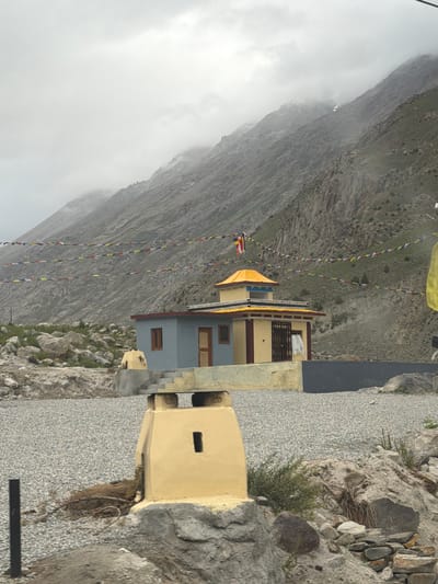 The image depicts a Buddhist monastery or stupa in Chikka, India, set against a backdrop of steep, rocky mountains partially obscured by clouds. Prayer flags are strung across the scene, a common sight in Buddhist regions. In the foreground, a yellow, cuboid structure with a small rectangular opening is visible. Behind this, a light blue and yellow building with a red-trimmed roof stands, featuring two windows and a door with a barred gate. The ground is covered in gravel, with scattered rocks and sparse vegetation. The atmosphere appears overcast and possibly damp, suggesting a cool climate. No people are present in the frame.