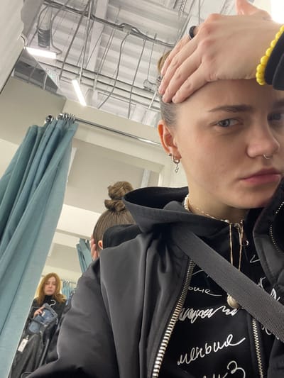 A selfie shows a person indoors, in a space with exposed pipes and fluorescent lighting. The person, with short hair and facial piercings, wears a black jacket with a black hooded sweatshirt underneath. A hand touches the forehead. In the background, draped light blue fabric is visible. Reflected in a mirror are two other people. One has their hair in a bun and is wearing a similar black jacket. The other has longer, lighter hair and wears a black coat while holding a blue bag.