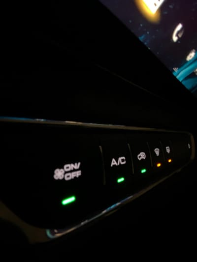 A close-up shot captures a car's climate control panel in the dark. Several buttons are visible, each labeled with a function such as "A/C" and "ON/OFF". Small indicator lights accompany each button, with some illuminated in green and others in orange. The control panel appears to be part of a modern car, with a sleek black finish and a subtle chrome trim. The image focuses sharply on the buttons and their corresponding lights.