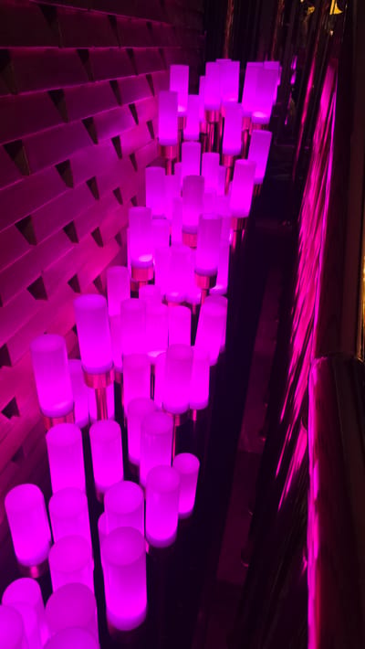 In Kazafani, Northern Cyprus, an indoor scene features a textured wall illuminated by vibrant pink lights. Extending from the floor are numerous cylindrical lamps, also glowing with pink light. These lamps are arranged in clusters and at varying heights, some with metallic bases. The floor appears to be dark and reflective, mirroring the light from the lamps. The overall ambiance is modern and artistic, with the interplay of light and shadow creating a dramatic effect.