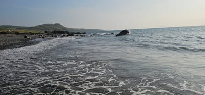 The image depicts a rocky beach with waves gently lapping at the shore. In the foreground, the water is foamy and turbulent, with a pattern of white froth on the dark surface. The beach itself is composed of dark pebbles and larger rocks. In the middle ground, larger rocks emerge from the water, and a prominent dark boulder stands against the incoming waves, with a splash of water visible. The background features rolling green hills that rise behind the coastline. The sky is a pale, clear blue, suggesting it is daytime. The scene is located on the coast of Girvan, United Kingdom. There are no people or animals visible in the image.
