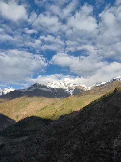 A dramatic mountain landscape is captured under a sky filled with fluffy white clouds against a bright blue backdrop. The sun casts long shadows across the rugged terrain, highlighting the green, grassy slopes in some areas while plunging others into deep shade. Snow-capped peaks rise majestically in the distance, their white summits contrasting with the darker, rocky mountain faces.
The time of day appears to be either early morning or late afternoon, judging by the angled sunlight and the presence of distinct shadows. The weather seems fair, with the clouds suggesting a partly sunny day. The rugged, mountainous terrain with patches of green vegetation and snow-covered peaks points to a location like Stingri, India, known for its high-altitude scenery.
The scene evokes a sense of awe and the grandeur of nature. There are no visible people or man-made objects, focusing the viewer's attention entirely on the natural environment. The composition emphasizes the scale and raw beauty of the mountains and sky. No text is visible in the image.