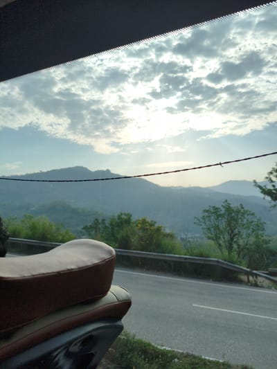 A medium shot captures a motorcycle seat in the foreground, viewed from the rear and side. The seat is brown with darker brown trim. Behind the motorcycle, a two-lane asphalt road curves to the right, bordered by a metal guardrail. Beyond the road, lush green hills and mountains recede into the distance, fading into a hazy blue. The sky above is filled with scattered white and grey clouds, with sunlight breaking through in rays, indicating it is likely early morning or late afternoon. A thin, dark wire stretches horizontally across the middle of the frame, obscuring part of the distant mountain range. The scene suggests a scenic route, possibly in the outskirts of Bilaspur, India, given the mountainous terrain and vegetation.