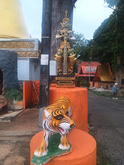 The image shows a scene in Ban Thung, Thailand. In the foreground is a painted tiger statue, orange with black stripes and white markings on its face and legs, positioned atop an orange, circular stand. Behind the tiger, an ornately decorated blue, gold, and white giant figure stands on an orange cylindrical base next to a grey utility pole with an electrical box affixed to it. To the left of the giant is a small building painted grey and beige with ornate gold details, while behind it sits a golden dome. In the background, there are several traditional Thai buildings with red roofs, orange walls, and golden accents. A person wearing a helmet and a light blue jacket is walking on a road, and there is some construction material scattered around. The sky is overcast.