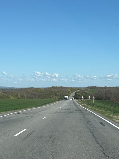The image shows a straight asphalt road in Pokrovskoye, Russia, flanked by a landscape of trees and green grass on one side and a clear blue sky with scattered clouds on the other. A few vehicles are seen driving on the road in the distance. Road markings, including white lines, are visible on the road surface.