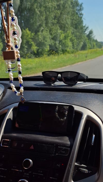 The image shows the interior of a car dashboard on a sunny day, viewed from the passenger seat. On the dashboard, a pair of black sunglasses rests on a textured surface. To the left, a beaded car accessory with a charm hangs from the rearview mirror, partially obscuring the view of the windshield. The accessory is made of white and blue beads with a small wooden cube and a cartoon character. A navigation screen is visible below the sunglasses, displaying a map. The car's central console with various buttons, including a "SPORT" and "TOUR" button, is also visible. Outside the car, through the windshield, a green, treed landscape and a road are visible under a clear blue sky. The overall impression is a snapshot of a car journey in a rural setting.