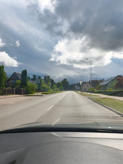 The image shows a view from inside a vehicle looking out at a paved road in a suburban or semi-rural area. The road stretches into the distance, with sidewalks and residential buildings lining both sides. There are trees and vegetation present. The sky is cloudy, with some dark, heavy clouds indicating potential for rain or stormy weather, contrasted with brighter areas. Streetlights are visible along the road. The dashboard of the vehicle is visible at the bottom of the frame. This scene is consistent with a street in Madona, Latvia, given the residential architecture and vegetation typical of the region. There are no people or specific actions depicted in the image.
