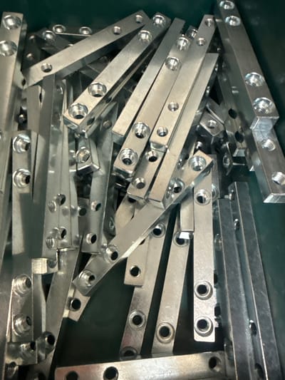 A pile of shiny, metallic, rectangular components are tightly packed together in a dark green container. These components appear to be manufactured metal parts, each featuring multiple circular holes, some of which are threaded. The lighting casts strong reflections off the metallic surfaces, creating bright highlights and deep shadows, obscuring some of the finer details of the pile. The composition is a close-up, overhead shot. This image was taken in Fontevivo, Italy.