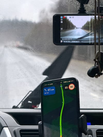 The image, taken from inside a vehicle looking through a wet windshield, depicts a journey during early evening in potentially rainy conditions. Water droplets and streaks are visible across the windshield, obscuring parts of the exterior view.
Two smartphones are mounted within the vehicle. The phone on the left, likely a dashcam or acting as one, displays a live recording of a straight road flanked by trees. Its screen shows a red recording indicator, various interface icons, and overlaid text: "AABCDEF", "19:04:19", and "2026/04/21". This indicates a recording timestamp of 7:04 PM on April 21, 2026. The view through this screen suggests an overcast sky.
The smartphone on the right displays a navigation application. A green route line is prominent on the map, with "E22" visible along the path. The current speed is shown as "85" (likely km/h), with a posted speed limit of "90" km/h. Below the speed information, the display indicates "41 KM" remaining and the road name "M-12". The phone's status bar shows a battery level of "51%" and the time "18:41" (6:41 PM).
The scene captures a person driving on the M-12 highway in Russia, as indicated by the navigation details and the provided location context of Kiselevka, Russia. The simultaneous use of a dashcam and navigation suggests active travel.