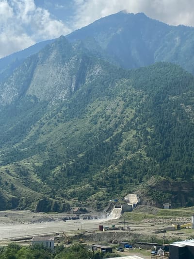 The image depicts a mountain landscape with a significant man-made structure, likely a hydroelectric dam, built into a steep hillside. Water cascades down the concrete spillway of the dam, flowing into a river or reservoir below. The surrounding mountains are covered in dense green vegetation, with a mix of trees and scrub. The sky above is partly cloudy. In the foreground, there is evidence of construction or industrial activity, including buildings, construction equipment such as a crane and excavators, and various storage containers. The overall scene suggests a remote, mountainous region where infrastructure has been developed to harness natural resources. The location is identified as Chalda, Russia, a village known for its proximity to natural landscapes and potential for hydropower development.