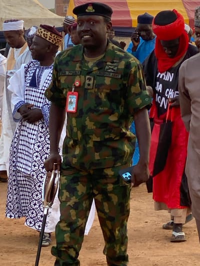 A smiling man in a Nigerian Army uniform and beret stands amongst others. He holds a ceremonial staff. His uniform features camouflage print, insignia, and an ID card. Others around him include men in traditional clothing, such as patterned robes and hats. A person wearing a red robe with black lettering is also visible. The event seems to be taking place outdoors on a sandy or dirt surface.