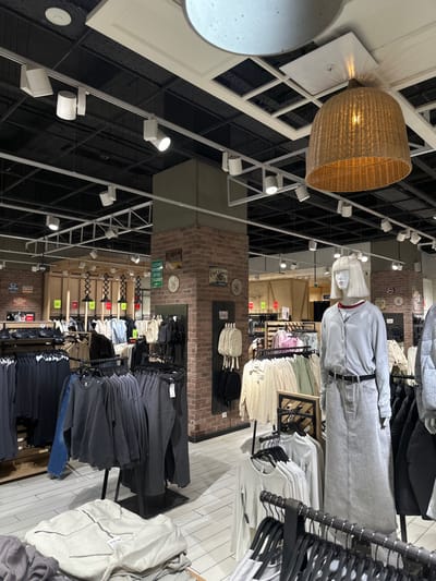 A department store interior features clothing racks filled with shirts and pants, including a mannequin dressed in a grey outfit with a belt. Brick pillars separate sections with assorted t-shirts and folded items displayed on shelves. The ceiling showcases industrial lighting, metal support beams, and a straw-like decorative fixture.