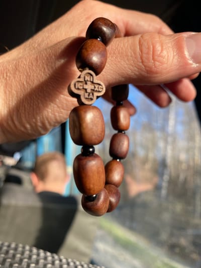 A person's hand holds a wooden bracelet with a cross charm. The bracelet is made of rounded, brown wooden beads, interspersed with small black beads. The cross charm is also made of wood and has a distinct design carved into it. The background shows a blurry interior, possibly a vehicle, with glimpses of someone's head and clothing. The location is not verifiable as Mishutino, Russia based solely on the image.