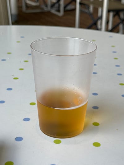 The scene depicts a single translucent plastic cup positioned centrally on a white table surface in an indoor setting within the city of Puerto del Rosario, Spain. The cup is approximately half-filled with a golden-amber liquid, consistent with beer, exhibiting a minimal amount of white foam at the liquid's surface and condensation on the cup's exterior. The table surface features a repeating pattern of uniformly spaced circular dots in alternating light blue and lime green colors. The background is out of focus, showing blurred, light-colored structural elements, likely parts of chairs or other furniture. No individuals are visible, and no discernible actions or interactions are taking place.