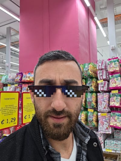 An adult male is depicted standing in a supermarket aisle located in Kato Polemidia Municipality, Cyprus. He has dark hair with visible grey, a short beard, and is wearing a white t-shirt beneath a dark, open jacket. His eyes are obscured by pixelated "Thug Life" style sunglasses as he looks directly forward. In the background, store shelves are visible, stocked with numerous brightly colored packaged confectionery items, including various sweets and snacks. To the left, two yellow price signs display "€ 1,29" and text in Greek, notably "ΓΛΕΙΤΖΟΥΡΙ" (Lollipop/Sucker). A large, bright pink structural pillar dominates the upper background, and overhead fluorescent lighting fixtures are present. The scene is brightly illuminated, characteristic of a commercial retail environment.