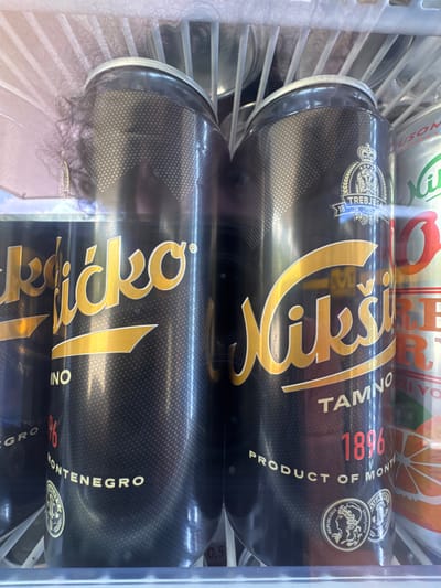 This image depicts the interior of a refrigerated display case in what is identified as a retail setting in Vrijesno, Montenegro. Two black aluminum beverage cans, prominently branded "Nikšićko Tamno," dominate the foreground. Each can features a textured dotted pattern, a gold stylized "Nikšićko" logo, and the word "TAMNO" (dark). The year "1896" is marked in red, accompanied by the text "PRODUCT OF MONTENEGRO." A royal-style crest with "TREBJESA NIKŠIĆ" and "1896" is positioned near the top of the can. At the base of the design, a gold circular medallion inscribed "REPUBLIQUE FRANCAISE" with a profile figure is visible. A third "Nikšićko" can is partially visible on the far left. To the right, a portion of a silver can with an orange graphic and red lettering ("USOM") is discernable. The cans are arranged on a white wire shelf typical of commercial refrigerators. Reflective surfaces show minimal condensation, and strands of dark, curly human hair are visible near the top left portion of the leftmost can, indicating close proximity or interaction.