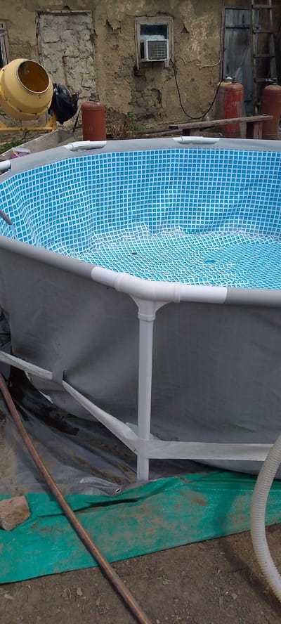 This image depicts an outdoor scene in Shkurinskaya, Russia, focusing on the construction or setup of an above-ground swimming pool. The pool itself is partially filled with water, revealing a blue tiled interior. White PVC pipes form the support structure for the pool's rim. The surrounding environment appears to be a yard or construction area, with a rough, textured building in the background. To the left, a yellow cement mixer and a black bag are visible. Red propane tanks are also present in the background. The ground is covered with a green tarp and dirt. A brown hose lies on the ground. The scene suggests an ongoing or recently completed project. No people are visible in the image.