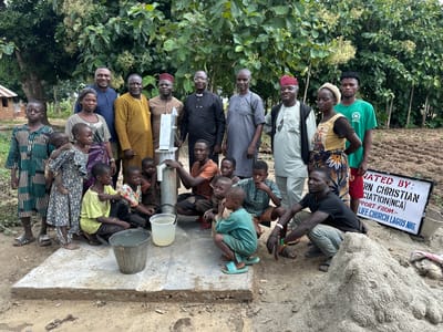 In Afogo, Nigeria, a group of approximately twenty people, comprising both adults and children, are gathered outdoors around a newly constructed water well. The individuals are of varying ages and genders, and many of them are dressed in traditional African attire. The scene appears to be a community event, possibly a dedication or unveiling of the well. The environment is rural, with a backdrop of trees and vegetation, and the ground is composed of dirt and some loose soil. A sign is visible in the background, which reads "RN CHRISTIAN ASSOCIATION (NCA) SUPPORT FROM:- LIFE CHURCH LAGOS NIG.". The people are interacting with each other in a relaxed and friendly manner, with some of the children playing near the well. The overall atmosphere is one of communal activity and shared purpose.