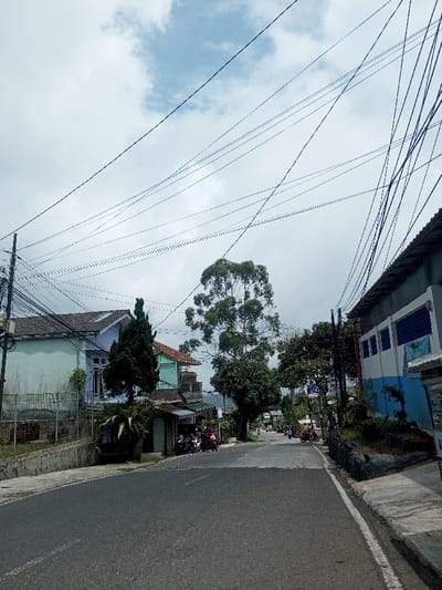 A street scene under a cloudy sky is depicted. Power lines crisscross overhead. Houses of varying colors line the left side of the street, while a building with blue accents stands to the right. Trees provide a canopy over the road further ahead. Several motorcycles are parked near a small shop, and a few vehicles are visible in the distance down the street. The road itself is asphalt, with a sidewalk running alongside the right side.