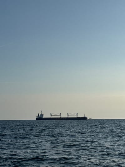 A large cargo ship is visible on the horizon, centered in the frame. The ship is dark and silhouetted against a pale sky. It has a superstructure at the rear and what appears to be cranes or cargo handling equipment along its deck. The ship is positioned on a dark blue, choppy sea. The sky is a gradient of light blue at the top fading to a lighter hue near the horizon, with no clouds visible. A smaller, indistinct object, possibly another vessel or a distant structure, is faintly visible to the right of the cargo ship on the horizon. The scene appears to be a wide, open body of water under a clear sky. The location is the Adriatic Sea, off the coast of Marina di Ravenna, Italy.