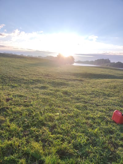 The image depicts a sun-drenched grassy field with a gentle slope towards a body of water. The sun is low on the horizon, casting a bright glare and creating long shadows. A red plastic container, possibly a gas can, rests on the grass in the lower right corner of the frame. The sky is a pale blue with scattered, wispy clouds illuminated by the sunlight. In the distance, a line of trees marks the opposite bank of the water. The overall scene suggests an outdoor setting in the early morning or late afternoon, with a focus on natural light and landscape elements. The location is identified as Архангель ауылдық округі, Kazakhstan.