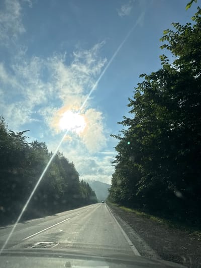 The image captures a view from the driver's perspective, looking out onto a paved highway that stretches into the distance. The road is bordered by dense, lush green trees on both sides. The sun is bright and high in the sky, with a strong lens flare diagonally crossing the scene from the upper left to the lower right. Wispy clouds are scattered across the blue sky, some illuminated by the sun, creating a bright halo effect around it. The road surface shows some wear, with patches and white lane markings visible. In the far distance, a small, light-colored vehicle can be seen on the highway, and beyond that, a misty mountainous landscape fades into the background. The overall impression is of a scenic drive through a heavily wooded, possibly mountainous, area.