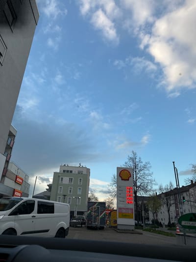 The image presents an eye-level view from inside a vehicle, likely a car or truck, focusing on a scene outside the driver's side. The photograph appears to have been taken in an urban setting, identified as Speyer, Germany, based on visible store signs and architectural styles.

The dominant feature in the image is a Shell gas station sign displaying current fuel prices. The prices are listed in Euros with varying decimals (e.g., 2.249 for Diesel).

Adjacent to the gas station, a "NORMA" discount grocery store is visible. A white van is parked near the store.

In the background, apartment buildings and other urban structures are present. The sky is partially cloudy. A traffic light signal is visible above the building.