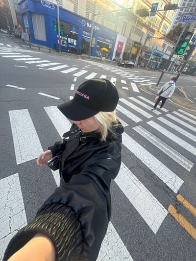 A person wearing a black leather jacket and a "Balenciaga" baseball cap stands on a crosswalk. The crosswalk features alternating black and white stripes. The person is looking down. Another person can be seen walking in the distance, also on the crosswalk. In the background, there are buildings, a street sign, and a traffic light showing a green walking symbol, suggesting it is safe to cross. The sun is shining.