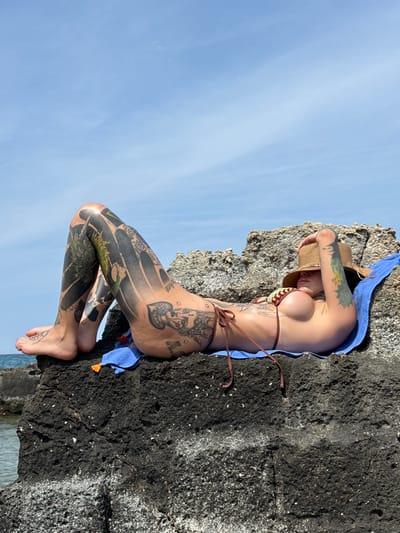 This full-shot image captures a person, likely female, lying horizontally on dark, textured coastal rocks. The image is presented in a portrait orientation, giving the impression the subject is positioned vertically on the rock. She is wearing a brown patterned bikini top and matching bottom, with a straw hat covering most of her face. A blue fabric, possibly a towel or sarong, is draped around her body, separating her from the rock in some areas.

The subject is extensively tattooed. Visible tattoos include: on her right arm, the English word "TATTOO" and Cyrillic text "Я НЕ КУРЮ" (meaning "I don't smoke"); on her hip, a large stylized face resembling a queen card with a crown, accompanied by the letters "A.N." and smaller text "chino". Her legs are almost entirely covered in intricate blackwork tattoos featuring green and white accents, such as a dragon design.

The setting is bright and sunny, with a clear, light blue sky dominating the left side of the frame. The rugged, dark rock formation takes up the right side of the image, showing signs of weathering and white speckled details. A small portion of calm water is visible at the bottom right, suggesting a seaside or coastal location. The overall atmosphere is one of relaxation, likely sunbathing on a warm day in Monopoli, Italy.