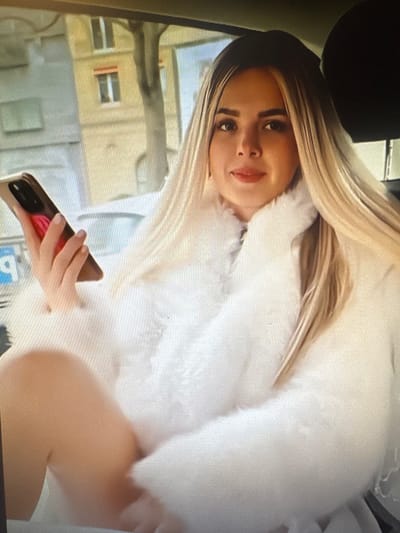 A young woman with blonde hair, wearing a white furry coat, sits in the back seat of a car. She is holding a smartphone in her right hand. The background suggests an urban environment with buildings and trees visible through the car window. It is not possible to determine the location in Chorzów, Poland from the image.