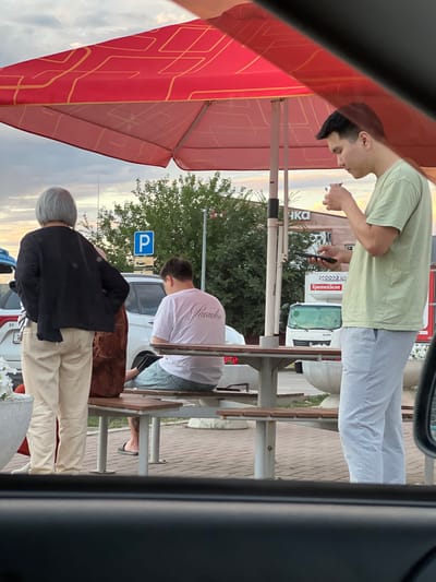 The image, taken in Zimovniki, Russia, shows three individuals in an outdoor setting, likely a cafe or rest area with outdoor seating. The time of day appears to be late afternoon or early evening, with a soft, warm light suggesting sunset.
In the foreground, a person with light gray hair and a black jacket is facing away from the camera, wearing light-colored pants. To their right, a person wearing a white t-shirt with the word "Paradise" printed on the back and light blue jeans is seated. They are looking down, and their legs are crossed. The third person, in the background, is wearing a light green t-shirt and light gray pants. They are also looking down and holding a phone in one hand and what appears to be a cigarette in the other.
The scene is set with tables and chairs, typical of an outdoor dining or resting area. A red awning with a yellow geometric pattern is visible on the left. In the background, there are trees, a white truck with Russian text that reads "Красное & Белое" (Red & White), and a parking sign with a blue "P". The ground is paved with bricks. The overall mood seems relaxed and casual.