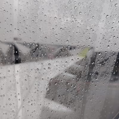 The image presents a close-up view through a transparent surface heavily coated with numerous water droplets of varying sizes. These clear raindrops refract and distort the out-of-focus background. The obscured background primarily consists of diffused white and light grey tones, indicative of a snow-covered or overcast outdoor environment. Indistinct, dark, vertical, and diagonal elements, consistent with bare tree branches or structural components, are vaguely discernible throughout the mid-ground. A subtle, diffused yellow-green tint is present in the upper-right quadrant of the background. No specific individuals, clearly defined objects, or discernible actions are visible beyond the environmental features. The scene is located in Skulte, Latvia, and suggests inclement, wet weather conditions.