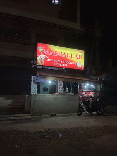 The image captures a nighttime street scene in B Camp, India, focused on a brightly lit "MASHALLAH MUTTON & CHICKEN CENTER" sign. The sign, predominantly yellow and red, features graphics of a rooster and a ram. Below the sign, a small shop or stall is visible, illuminated by artificial lights, with some food items or displays inside. A man is seated on a motorcycle parked in front of the stall. The street in the foreground appears to be unpaved or rough. The overall atmosphere is that of a commercial establishment operating at night in a somewhat rustic urban environment.
