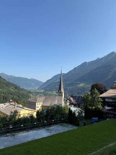 The image depicts a view of the town of Bad Gastein, Austria, on a clear day. In the foreground, a grassy slope leads down to a paved area and a green metal fence. Beyond the fence, several buildings are visible, including a prominent church with a tall, pointed steeple and a clock face. The church is situated in the center of the frame, surrounded by other structures with tiled roofs.
Further in the background, a picturesque valley opens up, framed by lush green mountains that are densely covered with trees. The mountains recede into the distance, with layers of them visible under a clear blue sky. The architectural style of the buildings suggests a traditional European alpine town. A dark blue van is parked to the right of the frame, partially obscured by trees. The scene conveys a sense of tranquility and natural beauty.