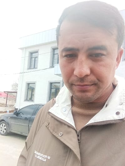 The image depicts an adult male taking a selfie outdoors in Nurabad, Uzbekistan. The subject, positioned in the foreground, has short dark hair, light facial stubble, and wears a light brown or khaki jacket with a white collar, silver snap buttons, and a visible tan turtleneck sweater underneath. Reversed text, appearing as "DR-PRODUCTIONS ALL-TECH", is present on the lower front of the jacket. Behind the subject, to the left, stands a two-story white building featuring multiple dark-framed rectangular windows and a small white balcony on the second floor. A dark-colored, visibly dusty sedan is parked on the light-colored ground between the subject and the building. In the distant background, faint structures and sparse vegetation are visible under an overcast, light grey sky.