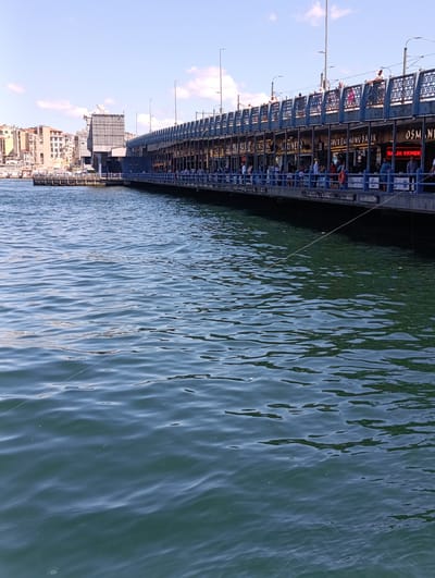 The image depicts the Galata Bridge in Fatih, Istanbul, Turkey, spanning across the Golden Horn. The bridge is lined with shops and restaurants, and numerous people are visible walking along the top and fishing from the lower level. The water below is a deep green, with ripples indicating slight movement. The sky is a clear blue with a few scattered clouds. The architecture of the bridge and the surrounding buildings suggest a bustling urban environment.