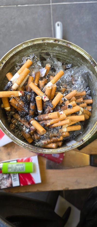 An overhead, close-up shot captures the interior of a large, round, metal receptacle filled with numerous cigarette butts. The butts are of various lengths and are predominantly orange and white, with some showing signs of ash and burns. The receptacle itself appears to be a repurposed tin can, with visible rust and wear on its rim. The surrounding environment suggests an outdoor setting, with a textured grey surface, possibly a tiled patio, visible in the upper portion of the image. A white object, potentially a handle or a light fixture, is positioned in the background. In the foreground, a green lighter rests on top of a pack of cigarettes, which features red and white graphics and Korean text. The overall scene is dimly lit, with the focus sharply on the collection of cigarette butts within the receptacle.