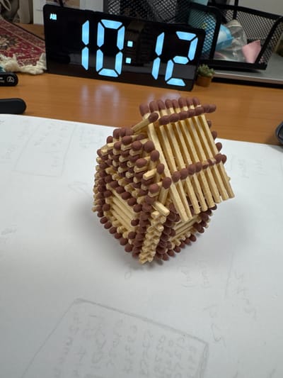 The image shows a close-up, high-angle view of a structure made of matches, placed on a white surface that appears to be paper with faint writing. To the left, a digital alarm clock displays "10:12 AM". In the background, a wooden desk is visible, with a rug and a black mesh organizer containing various items. The lighting is bright, suggesting an indoor setting. The scene does not contain any information that would allow for the identification of the city of Karmiel, Israel, or any specific location within it.