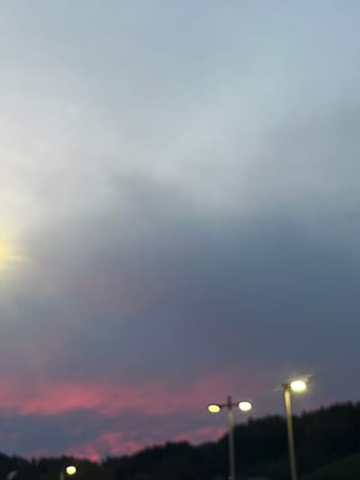 The image displays an outdoor scene in Oppenau, Germany, at either dusk or dawn. The sky presents a gradient from a bright, diffuse light source in the upper left, transitioning to grey, blue, and purple tones, with a prominent horizontal band of vivid pink and magenta clouds across the lower portion. Below the cloud line, a dark, silhouetted landscape of forested hills is visible. In the lower frame, three streetlights are illuminated. Two streetlights featuring dual circular, yellow-white fixtures are located right of center, while a third single streetlight with a similar fixture is partially visible at the bottom left. No persons or activities are present.