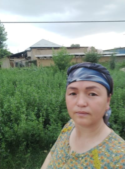 The image, taken in the city of Маданият, Kyrgyzstan, captures a frontal medium shot of a woman in her 40s with fair skin, brown eyes, and dark hair. She is wearing a blue and white patterned headscarf and a floral-print blouse in shades of green, yellow, and orange. The woman is positioned in the foreground, with a backdrop of lush green foliage and buildings. The background shows a cluster of residential structures with various roof styles, suggesting a rural or semi-urban setting. The sky is overcast with clouds, casting a diffused light over the scene. A thin wire is visible stretched horizontally across the upper portion of the image. The overall impression is that of an outdoor portrait taken in a residential area.