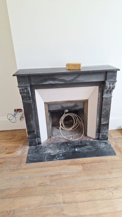 The image shows a fireplace in a room with light-colored walls and wooden flooring. The fireplace is made of dark stone with ornate carvings on the sides and mantelpiece. A light-colored coiled cable is placed inside the fireplace opening. To the left of the fireplace, electrical wires and conduits are visible, connected to an outlet on the wall. A small, rectangular object that appears to be a piece of foam or a sponge rests on the mantelpiece. The room appears to be undergoing renovation or is in a state of disarray, indicated by the exposed wiring and the general unfinished appearance of the area around the fireplace. The flooring is composed of wide, light-brown wooden planks. The scene does not provide any specific visual cues that would allow for precise localization to the city of Fontainebleau, France.