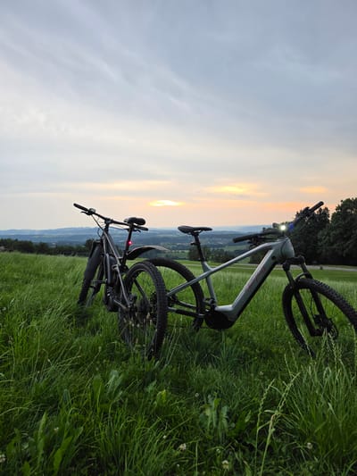 The image depicts two electric bicycles parked on a grassy hillside overlooking a rural landscape at dusk. The sky is overcast with streaks of pale orange and yellow near the horizon, indicating sunset. The foreground is dominated by tall, green grass. The bicycle on the left is dark gray and appears to be a mountain bike. It has a red taillight illuminated. The bicycle on the right is lighter gray and also appears to be an electric mountain bike, with its headlight on. Both bicycles are positioned at an angle, facing away from the viewer. In the background, rolling hills and scattered trees are visible, with a small town or village nestled in the valley. The overall scene suggests an outdoor recreational setting in a peaceful, rural environment. Based on the provided context, this scene is located in the vicinity of Bernhardzell, Switzerland. There are no people present in the image.
